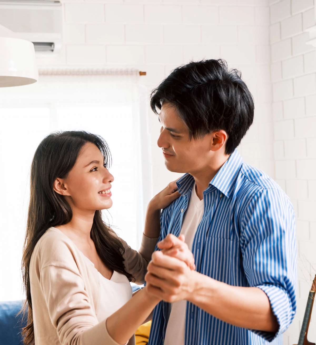 A young couple holds hands and dances together indoors, looking at each other and smiling in a bright, modern room.