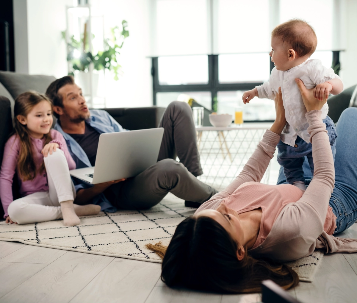 A family spends time together in a living room; a woman lies on the floor holding a baby, a man uses a laptop, and a young girl sits nearby, all appearing relaxed.