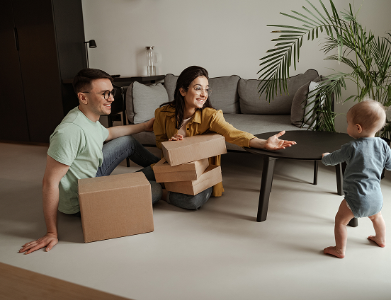 A man and woman sitting on the floor with cardboard boxes reach out to a toddler who is walking towards them in a living room.