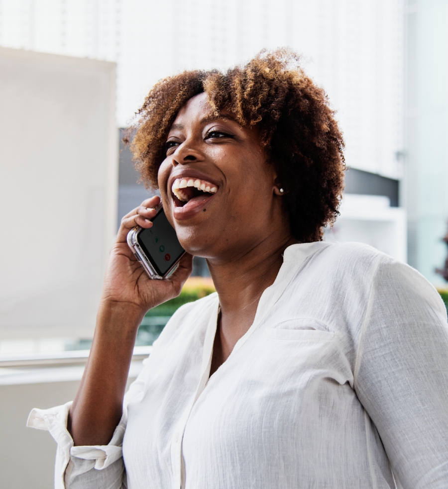 A woman wearing a white shirt is smiling and talking on a smartphone indoors.