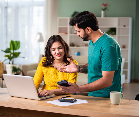 A woman using a laptop sits at a table while a man shows her something on his phone; papers, a calculator, and a coffee mug are on the table.