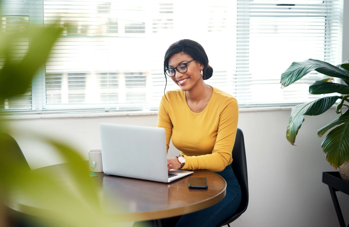 A woman wearing glasses and a yellow top works on a laptop at a round table in a bright office with large windows and plants.