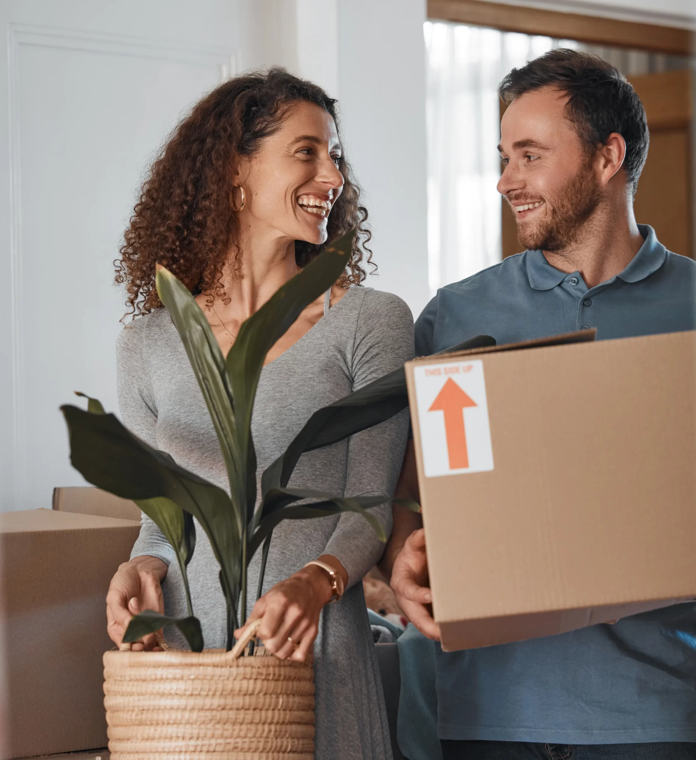A woman holding a potted plant and a man carrying a cardboard box smile at each other while moving into a home.