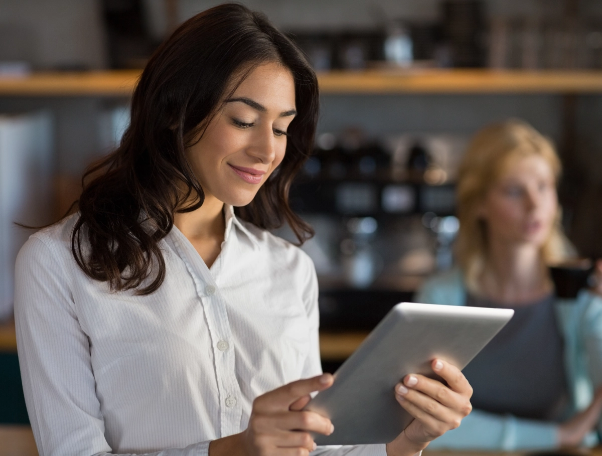 A woman wearing a white blouse is using a tablet while standing indoors; another person is blurred in the background.