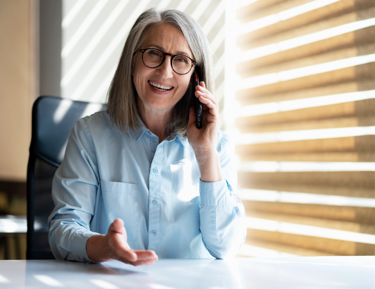 An older woman with gray hair and glasses, wearing a light blue shirt, sits at a desk smiling and talking on a mobile phone. Sunlight filters through window blinds behind her.