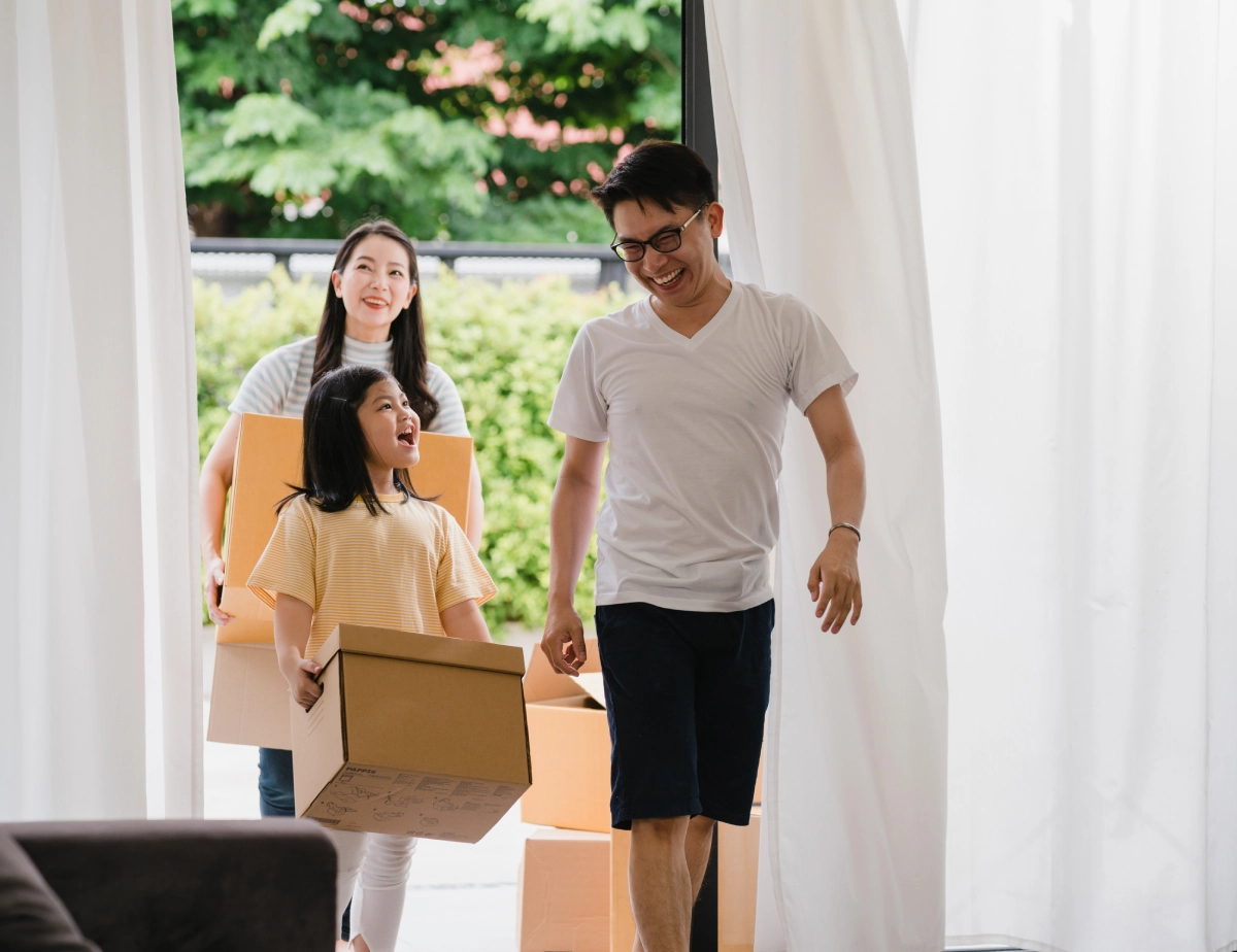 A man, woman, and young girl carry cardboard boxes into a bright room, appearing to move into a new home.