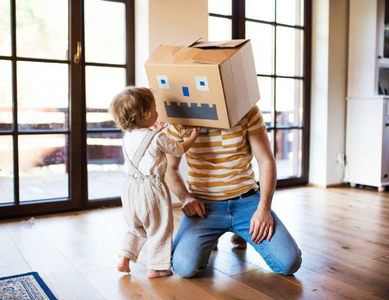 An adult kneeling on the floor wears a cardboard box with a robot face, while a toddler in overalls stands in front of them in a sunlit room with large windows.