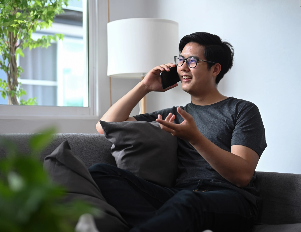 A person wearing glasses sits on a gray couch, talking on a smartphone and gesturing with one hand, with a lamp and window in the background.