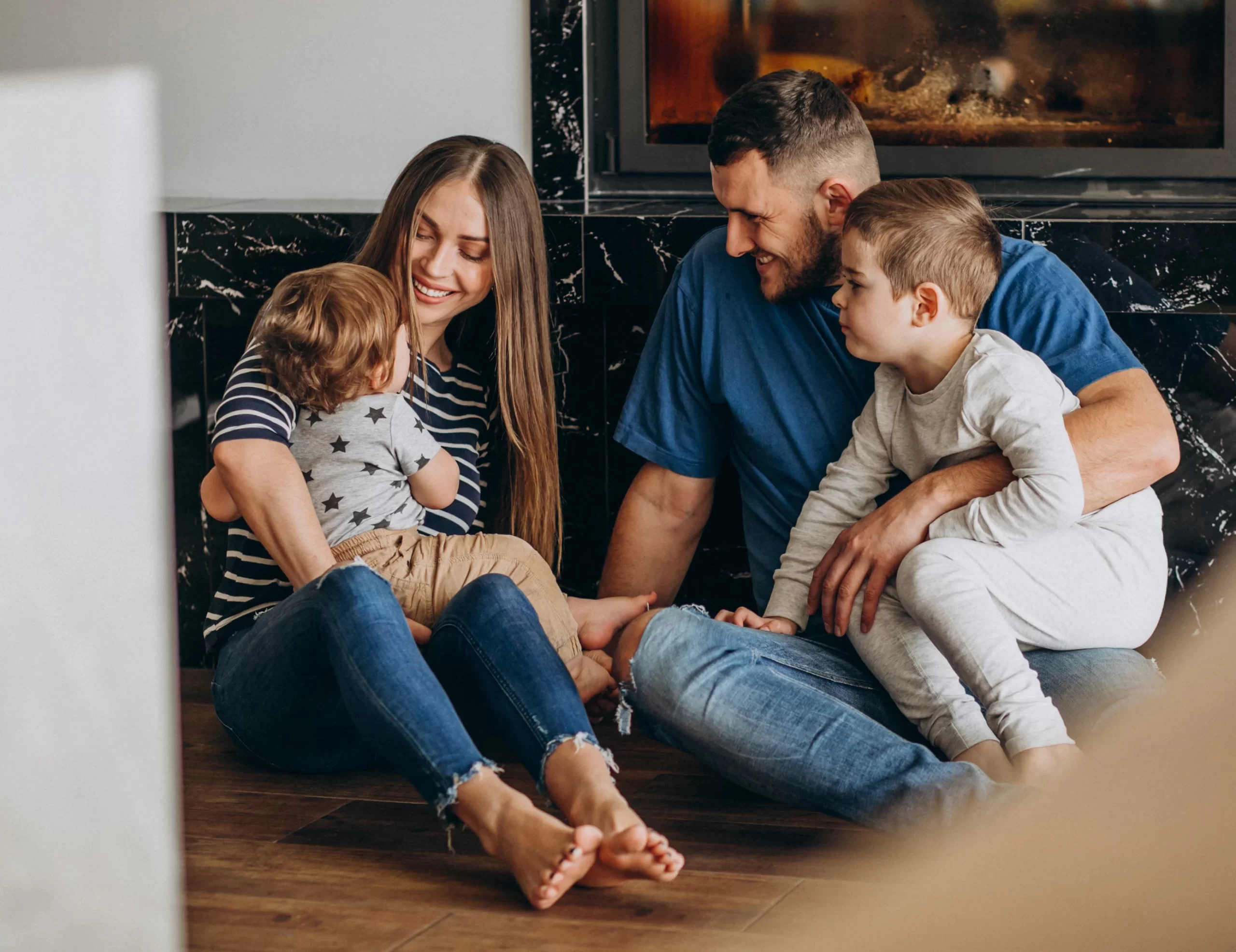 A family of four sits on the floor by a fireplace, with the parents smiling and holding their two young children.