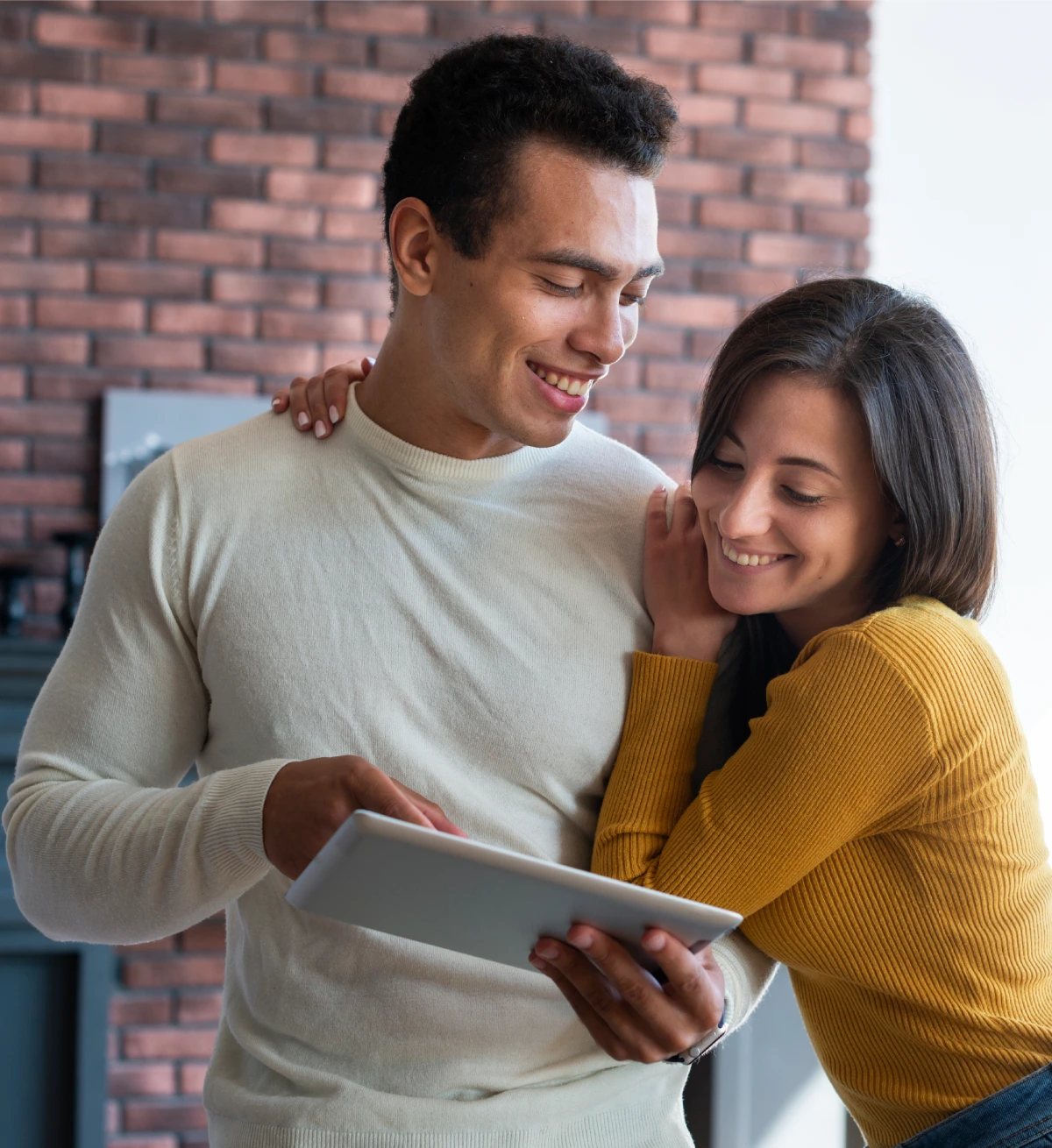 A man and woman standing close together, smiling while looking at a digital tablet indoors.