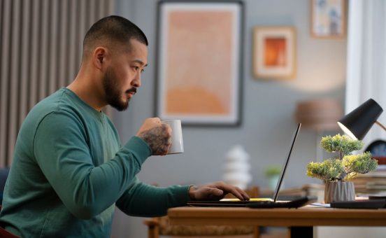 A man sits at a desk using a laptop and holding a mug, with a small potted plant and a desk lamp nearby in a home office setting.