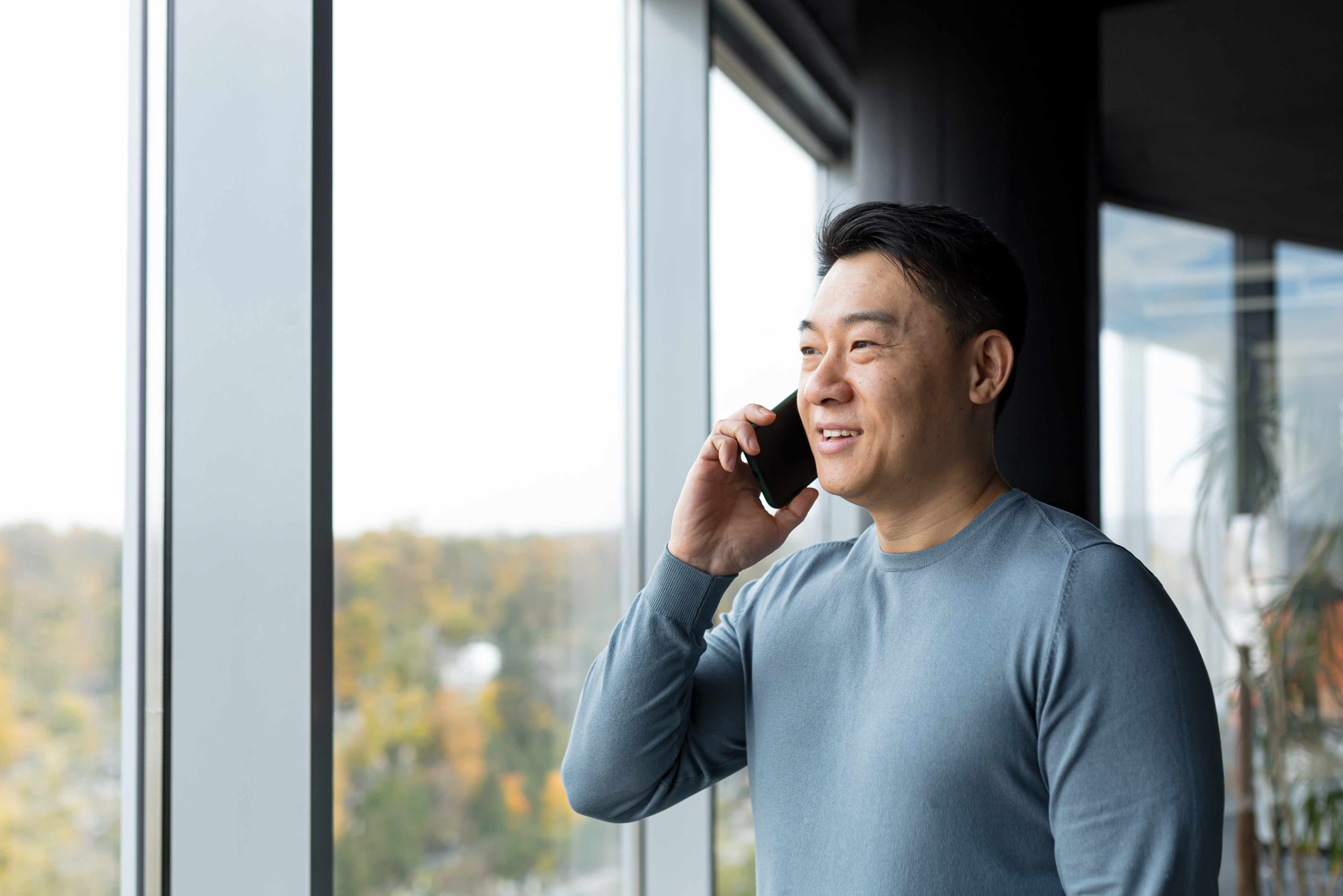A man stands by a large window, smiling while talking on a smartphone. Trees and sky are visible outside.
