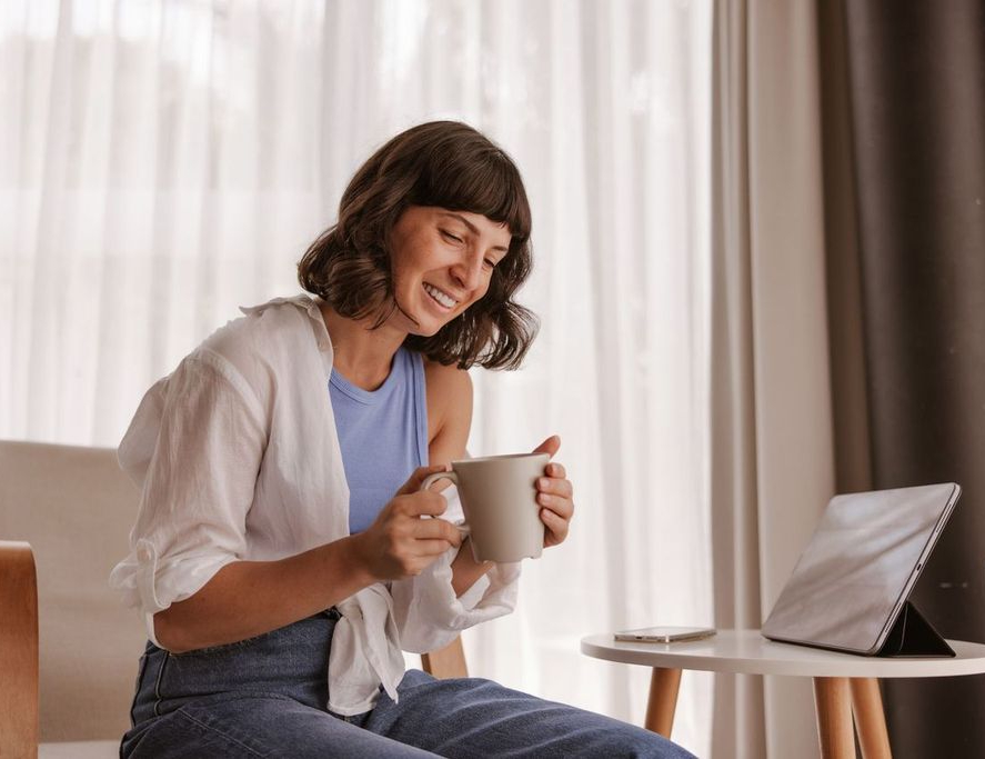 Woman sitting indoors, smiling while holding a mug and looking at a tablet on a small table, with a smartphone nearby.
