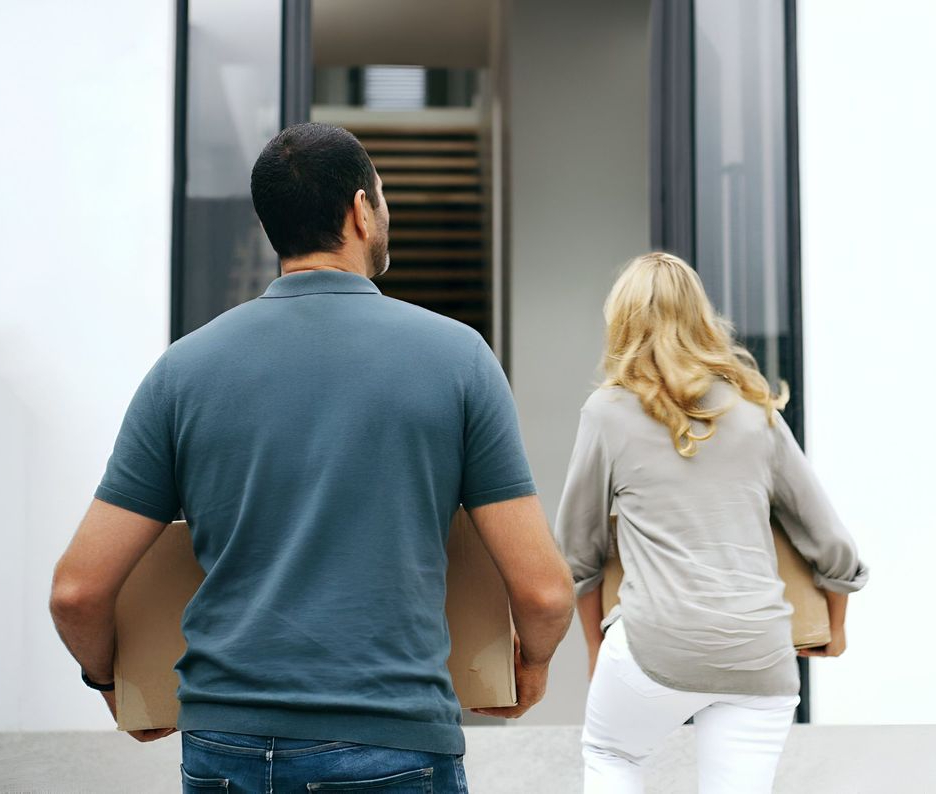 A man and a woman carrying cardboard boxes walk toward the entrance of a modern building.