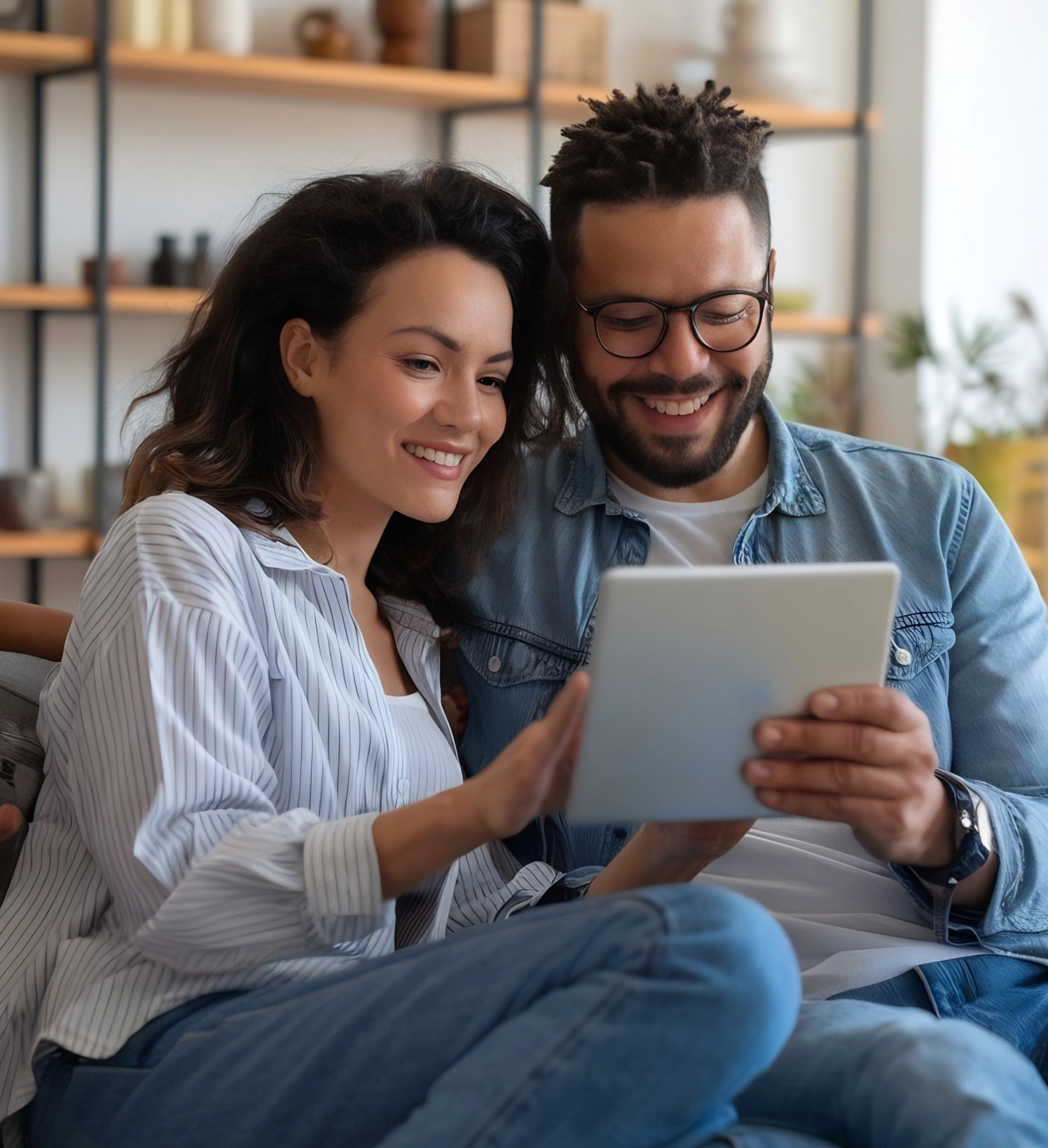 A woman and a man sit together on a couch, smiling and looking at a tablet device in a living room with shelves in the background.