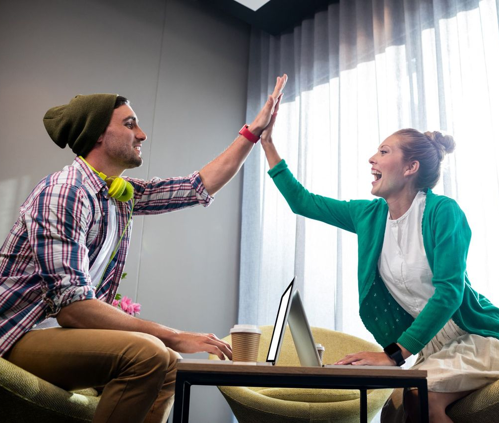 Two people sitting across a table high-five each other, smiling. A laptop, coffee cup, and papers are on the table between them.
