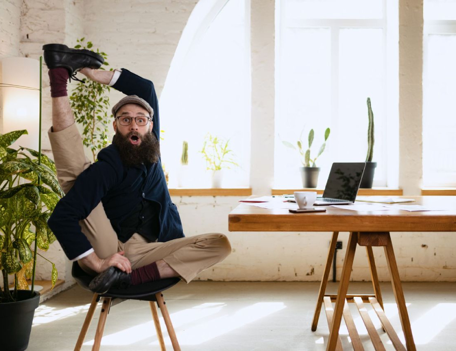 A man wearing glasses and a hat sits at a desk in an office, raising one leg high above his head while looking surprised.