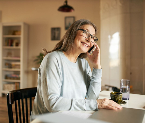 An older woman with long gray hair and glasses sits at a table, smiling while talking on a smartphone. A cup, glass of water, and papers are on the table.