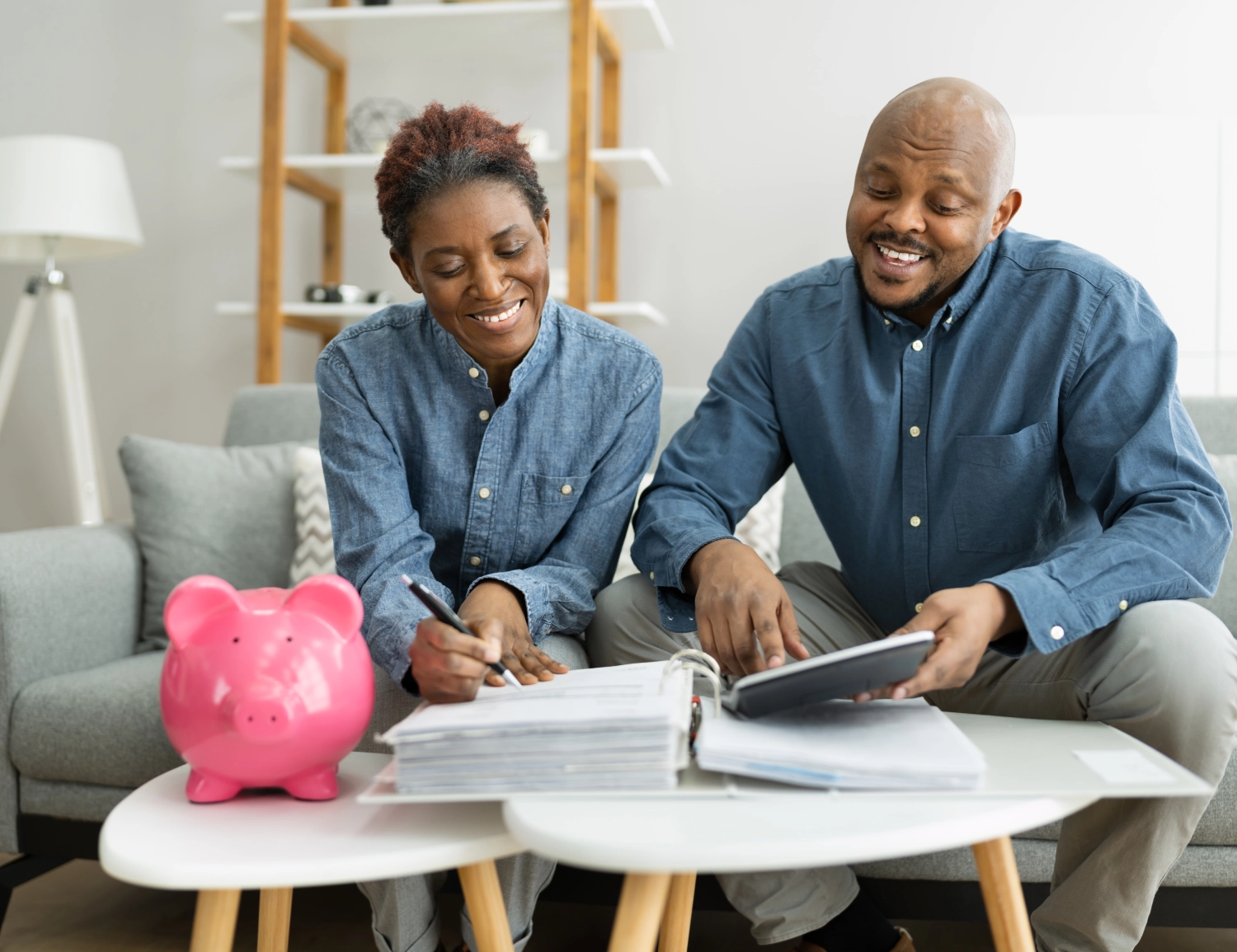 Two people sit at a table with a pink piggy bank, calculator, and paperwork, smiling as they review their finances together in a living room.