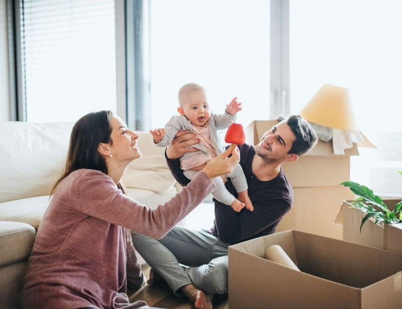 A woman and man sit on the floor with a baby, surrounded by moving boxes. The woman holds up a red toy for the baby.