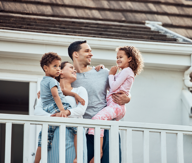 A family of four stands on a porch, with parents holding their two young children, smiling and looking at each other.