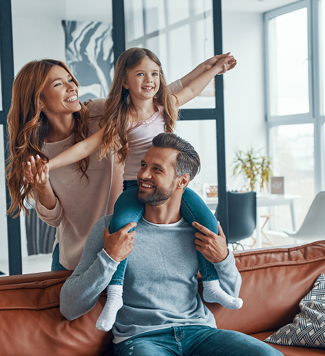 A man sits on a couch with a young girl on his shoulders and a woman standing beside them, all smiling in a bright, modern living room.
