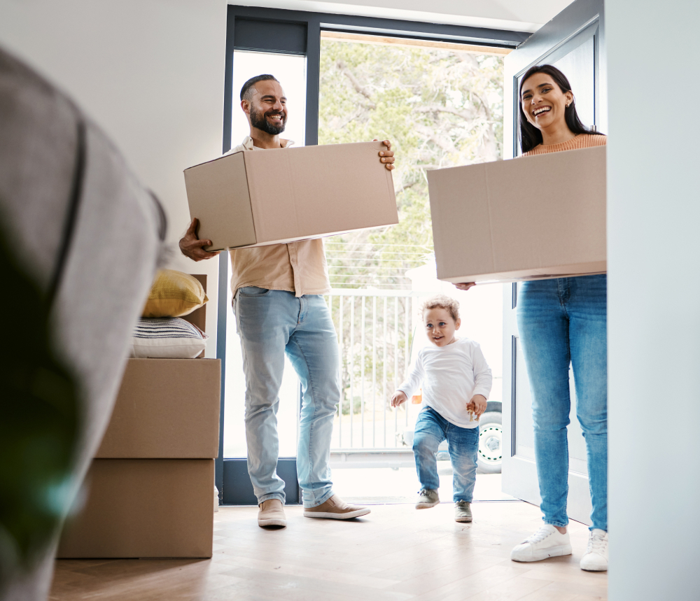 A man and woman carrying cardboard boxes enter a home, with a young child walking between them. More packed boxes are stacked nearby.