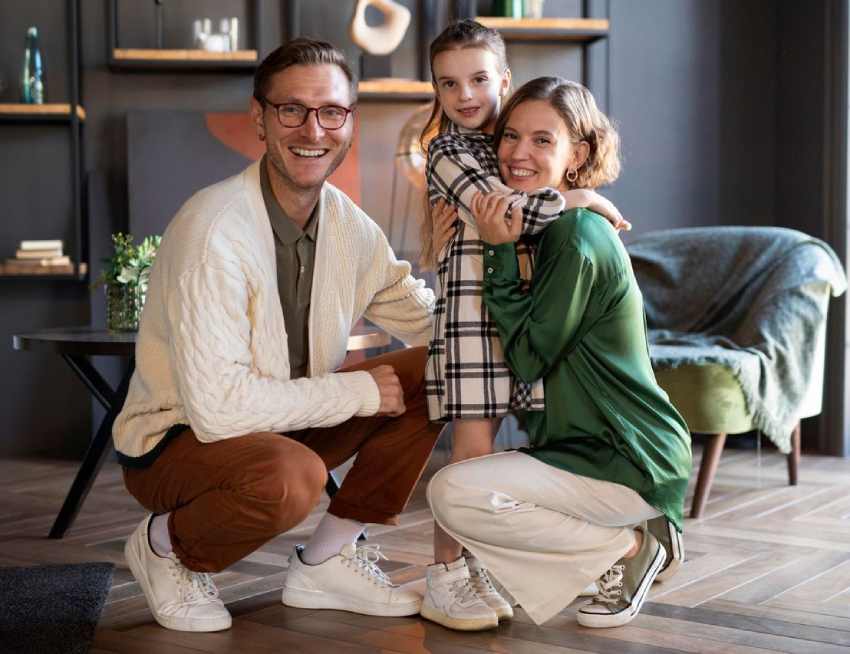 A man and woman crouch and smile with a young girl standing between them in a modern living room.