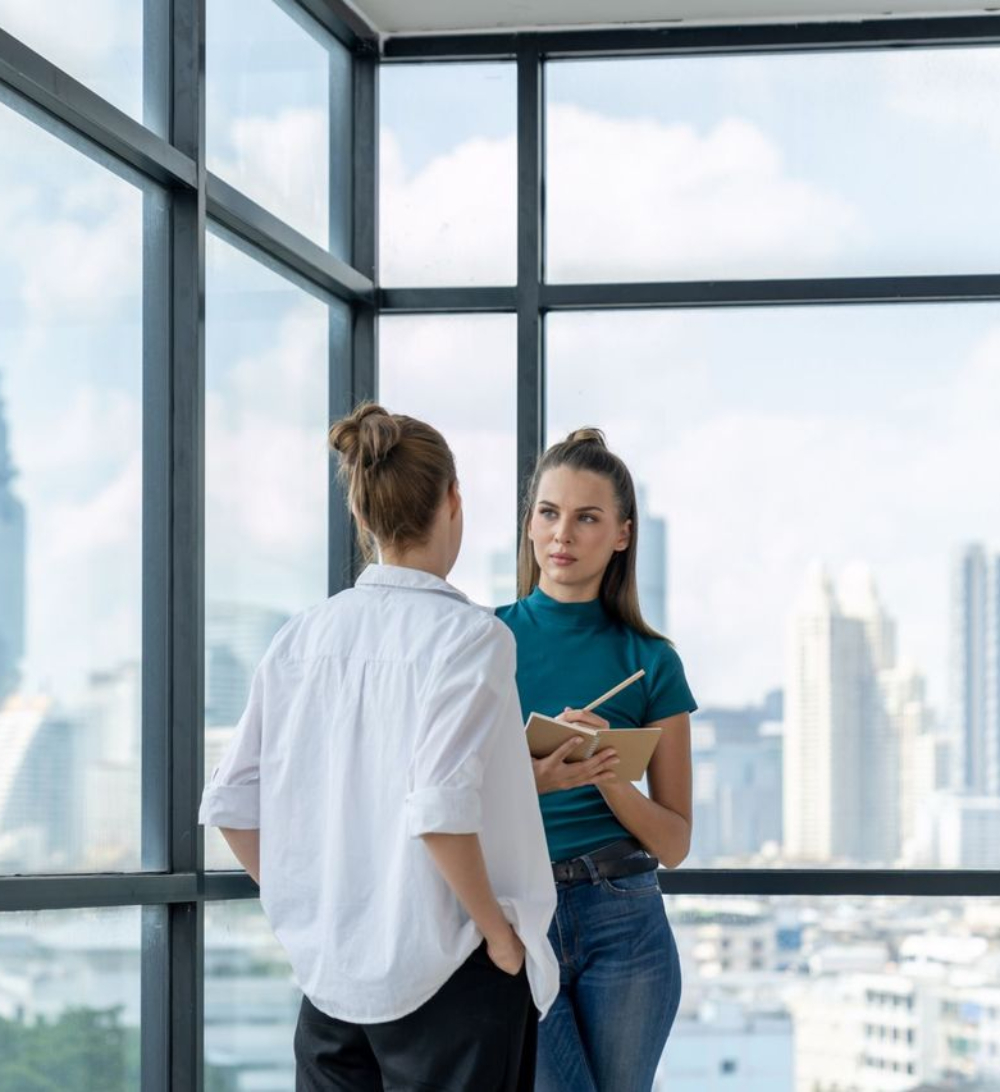 Two women stand by large windows in a modern office, one holding a notebook and pen, engaged in conversation with a cityscape in the background.