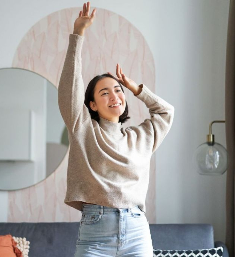 A woman in a beige sweater and light jeans stands indoors with one arm raised, smiling, in front of a circular wall mirror and patterned background.