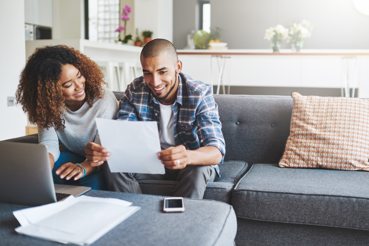 A man and woman sit on a couch, smiling while looking at paperwork; a laptop, phone, and more papers are on the table in front of them in a bright living room.
