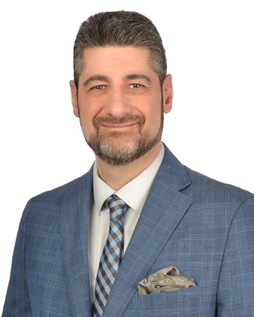 Man wearing a blue plaid suit, patterned tie, and pocket square, smiling at the camera against a white background.