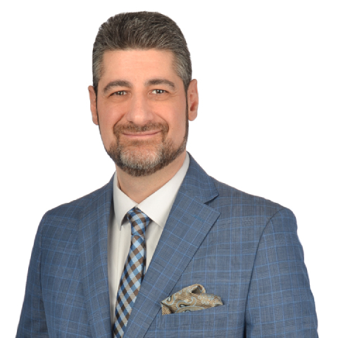 Man with short dark hair and beard wearing a blue checkered suit, patterned tie, and pocket square, smiling in front of a white background.