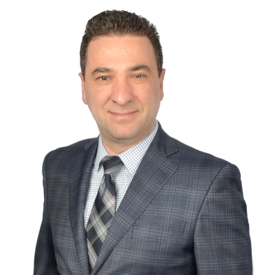 Man in a checked blue suit, patterned tie, and checked shirt, smiling at the camera against a plain white background.