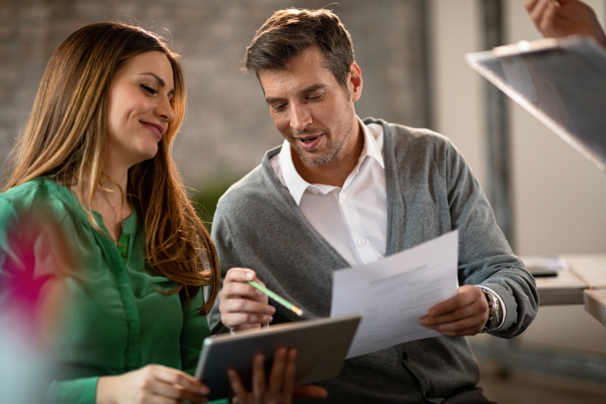 A man and woman sit together at a desk, looking at a tablet and paperwork, discussing something in an office setting.